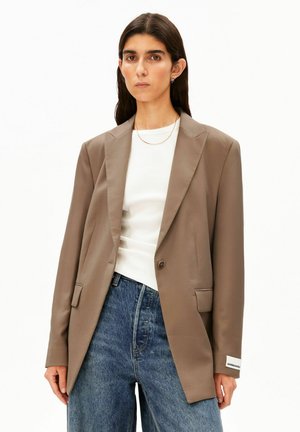 Vrouw draagt een taupekleurige oversized blazer, wit geribbeld topje en hoog getailleerde blauwe spijkerbroek, staand tegen een effen witte achtergrond.