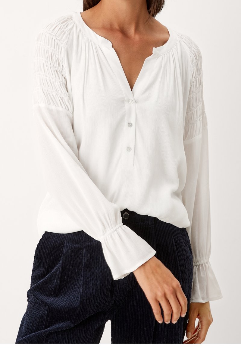 Camicia bianca a maniche lunghe con scollatura a V abbottonata, con maniche arricciate e una texture leggera, abbinata a pantaloni in velluto a coste scuri.
