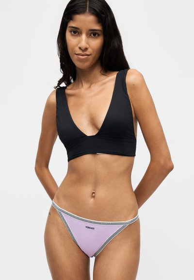 Femme aux longs cheveux foncés portant un haut de bikini noir à profond décolleté en V et un bas de bikini Versace lavande avec bord blanc et logo noir.