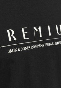 Juodas medvilnės T-shirt su ryškiai baltais užrašais, kuriuose parašyta „PREMIUM JACK & JONES COMPANY ESTABLISHED“, su lygiu, lengvu tekstūromis.