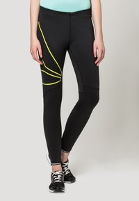 Schwarze Sportleggings mit gelben Akzentlinien, die ein figurbetontes Design und eine glatte Textur aufweisen. Gestylt mit schwarzen Sportschuhen.