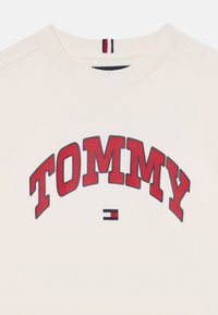 Lys kremfarget sweatshirt med en rød brodert "TOMMY"-logo, rund tekst, og en liten blå, rød og hvit stripe ved halsen.
