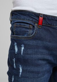 Acercamiento de unos pantalones de mezclilla desgastados de color azul oscuro con costuras naranjas y una etiqueta roja en el lazo del cinturón, combinados con una blusa blanca de canalé.