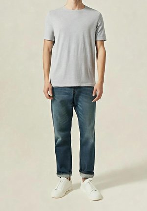 Homme debout portant un t-shirt gris clair à manches courtes, un jean bleu foncé remonté et des baskets blanches sur un fond uni.
