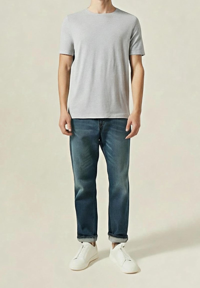 Homme debout portant un t-shirt gris clair à manches courtes, un jean bleu foncé remonté et des baskets blanches sur un fond uni.