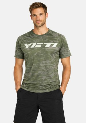 Uomo che indossa una maglietta YETI verde con motivo mimetico e pantaloncini neri, in piedi con le mani nelle tasche contro uno sfondo bianco.