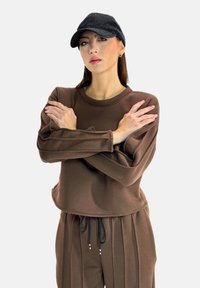 Bruine cropped sweatshirt gemaakt van zachte stof met lange mouwen; het model draagt het met bijpassende broek met verticale strepen. Een zwarte pet maakt de outfit compleet.