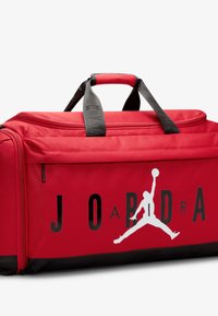 Piros sporttáska fekete részletekkel, tartós szövetből készült. Figyelemfelkeltő fehér Jumpman logóval és fekete felirattal, amely "JORDAN"-t ír.