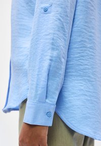 Μανσέτα γαλάζιου μακρυμάνικου πουκάμισου με κουμπί, φορεμένη από άτομο με σκούρο δέρμα, σε συνδυασμό με ανοιχτό ελιά παντελόνι, που δείχνει την υφή του υφάσματος και τις ραφές.