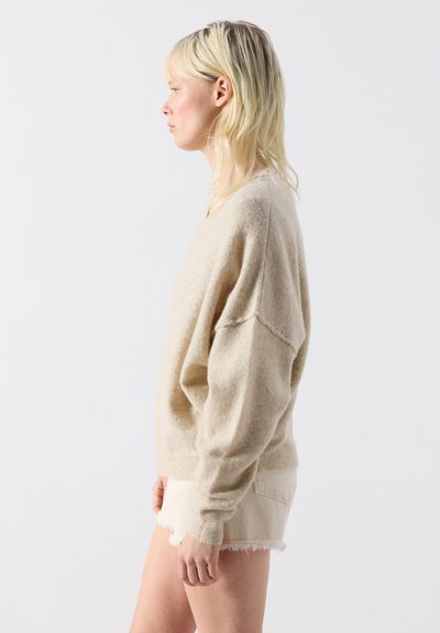 Femme blonde portant un pull beige oversize et un short clair effiloché, tournée vers la gauche sur un fond blanc uni.