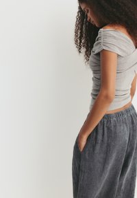 Kvinna medlockigt hår, iklädd en grå rynkad off-shoulder topp och lösa mörkgrå byxor med händerna i fickorna, vänd åt sidan.