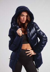 Mørkeblå pufferjakke med en blank finish, oversized hætte og frontlynlås. Har et logo-patch på ærmet og struktureret panelering.