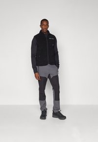 Μαύρο γιλέκο από φλις με μπροστινό φερμουάρ, υφή επιφάνειας και τσέπη με έμπλαστρο. Συνδυασμένο με γκρι παντελόνι που διαθέτει σχέδιο χρωματικών μπλοκ και μαύρα υποδήματα.