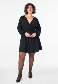 Melna iesaiņojamā kleita ar garām piedurknēm, dziļu V veida izgriezumu, savāktu vidukli un plūstošu svārku daļu. Stilizēta ar caurspīdīgām melnām zeķubiksēm un brūniem augstpapēžu apaviem.