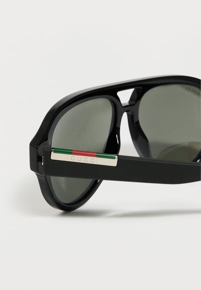 Γυαλιά ηλίου με μαύρο πλαίσιο και φακούς με πράσινη απόχρωση, που διαθέτουν χρυσή λεπτομέρεια με λογότυπο Gucci σε ρίγες κόκκινο-πράσινο στο πλαϊνό σκέλος.