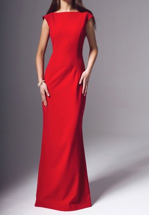 Femme debout, mains près des hanches, portant une robe rouge ajustée longue jusqu'au sol et un bracelet de perles sur un fond gris uni.