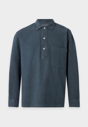 Mann trägt ein dunkelblaues Langarmhemd, beige weite Hosen und gelbe Schuhe und steht vor einem einfarbigen hellen Hintergrund.