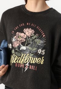 Sudadera negra de algodón con estampado gráfico floral en rosa y verde, con texto "Wildflower Rock & Roll" en amarillo, textura desgastada.