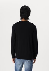 Suéter negro de manga larga con cuello redondo, puños y dobladillo acanalados, hecho de una tela suave. Visto desde la parte de atrás, combinado con jeans de un azul claro.