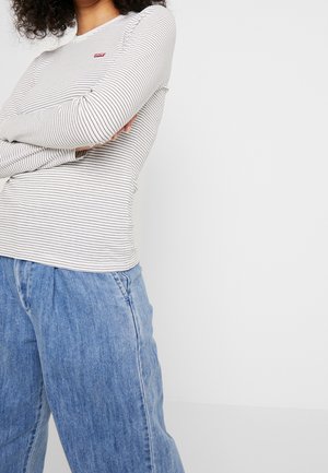 Person trägt ein weißes Langarmshirt mit dünnen schwarzen Streifen und blaue Jeans, die Arme verschränkt, vor einem schlichten Hintergrund.