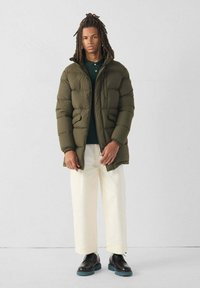 Olijfgroene pufferjack met een capuchon, voorzien van een gequilt ontwerp en zijzakken. Draagt over een donkergroen shirt en gecombineerd met crèmekleurige broek en zwarte schoenen.