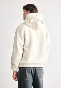 Hanorac alb, pufos, din fleece, cu guler înalt, având două urechi mici pe glugă. Asortat cu o pereche de blugi largi din denim, care prezintă o croială relaxată.