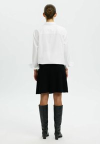 Balts garām piedurknēm krekls ar aizmugurē ievērstu piegriezumu, saskaņots ar melnu, platām apakšmalām svārku no teksturēta auduma un ceļa augstuma melnām ādas zābakiem.