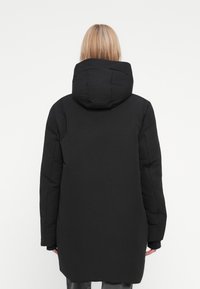Svart puffjakke med hette, som når midt på låret. Har et teksturert stoff, lange ermer og en avslappet silhuett.