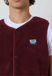 Burgundijas krāsas flīsa veste ar strukturētu virsmu, noapaļotu apkakli, priekšējiem pogām un krāsainu etiķeti ar uzrakstu "AMV SUPER VINTAGE."