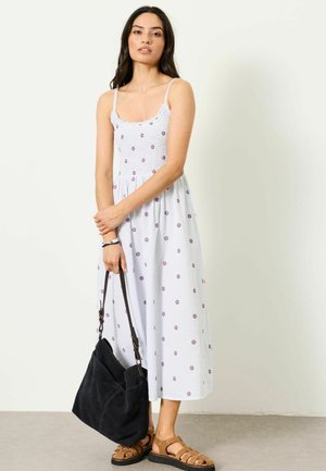 Sieviete baltā, bezroku midi kleitā ar maziem ziedu rakstiem, tur melnu plecu somu un valkā brūnus sandales.