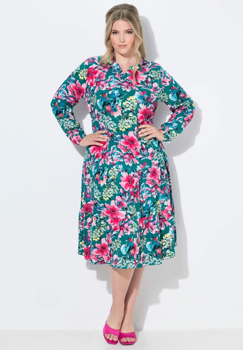 Vestido de estampado floral con mangas largas, fondo azul verdoso, flores rosas y verdes, cintura ajustada y falda acampanada. Combinado con zapatos de cuña rosas.