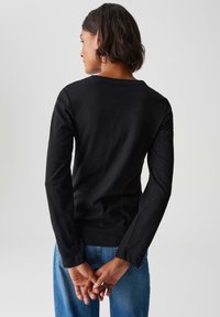 Frau mit kurzem dunklem Haar, die ein schwarzes Langarmshirt und eine blaue Jeans trägt, steht mit hinter dem Rücken gefalteten Händen.