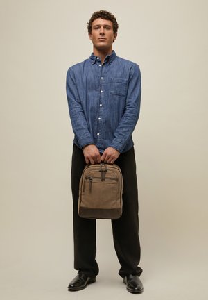 Muž stojí a drží béžový batoh oběma rukama, má na sobě modrou košili na knoflíky, černé kalhoty a černé boty na jednobarevném pozadí.