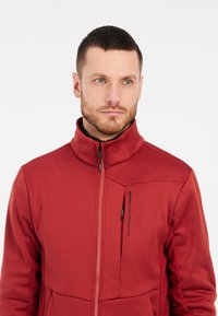 Rode softshell jas met hoge kraag, ritssluiting aan de voorkant en zwarte accenten. Heeft een gestructureerd patroon op de zijpanelen.