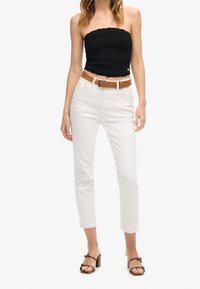 Woman wearing a black strapless top, white high-waisted pants with a brown belt, and brown heeled sandals, standing against a white background.