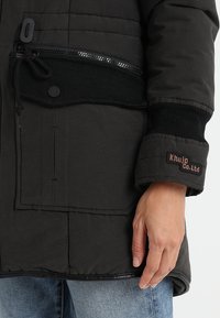 Giacca nera trapuntata con tasca con zip, polsini a coste e un patch con "Khujo Co. Ltd" sulla manica. Vestibilità casual e tessuto testurizzato.