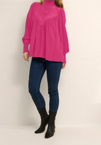 Fuchsia, lös passande blus med hög krage och voluminösa ärmar, ihop med mörkblå skinny jeans och svarta ankelstövlar.