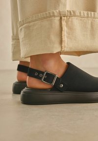 Mustat slingback-kengät, joissa on sileä materiaali, neliömäinen solki ja paksu pohja, valoisan tekstuurin päällä.