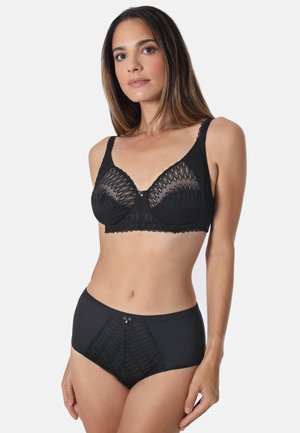 Frau trägt schwarzen Spitzen-BH und passende hoch taillierte Spitzen-Unterwäsche, steht vor einfachem hellem Hintergrund und schaut nach vorne.
