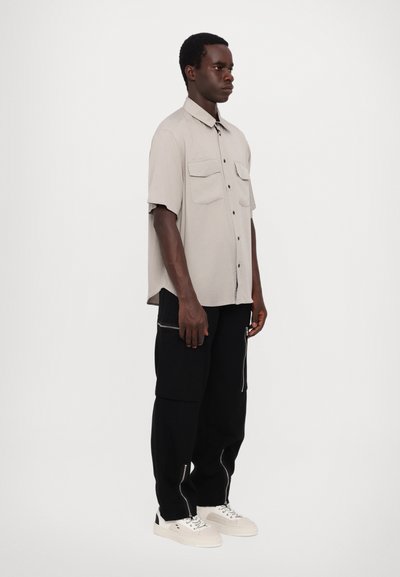Chemise beige à manche courte avec deux poches poitrine, associée à un pantalon noir avec des zips et des baskets blanches. Arrière-plan uni.