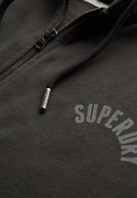 Grau zip-up Hoodie aus weichem Stoff mit schwarzen Kordeln und einem metallischen Reißverschluss. "SUPERDRY" ist in einer geschwungenen Schriftart gedruckt.