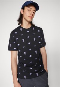 Camiseta de algodón negra con un logotipo de Nike repetido y texto "Just Do It" en morado. Cuello redondo y mangas cortas.