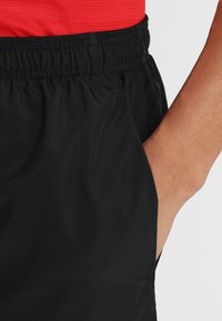 Pantalones cortos deportivos negros con cintura elástica, bolsillos laterales y una textura suave. Se ve una camisa roja encima de los pantalones cortos.