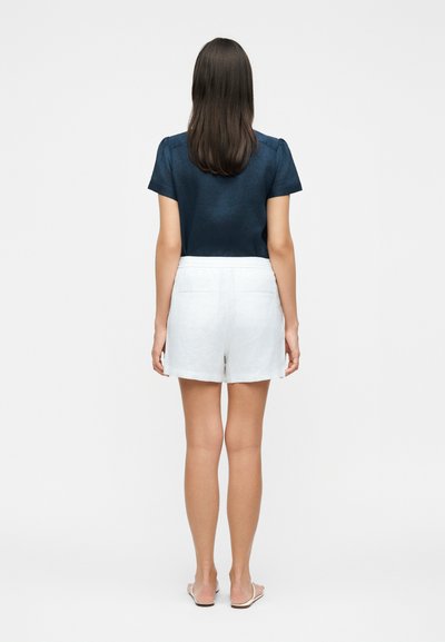 Γυναίκα που στέκεται γυρισμένη με την πλάτη, φορούν μπλε σκούρο κοντομάνικο τοπ, λευκά σορτς με πίσω τσέπες και λευκά σανδάλια σε απλό φόντο.