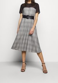 Robe à carreaux noir et blanc avec des manches courtes, un corsage ajusté et une ceinture noire à la taille. Couche supérieure transparente, silhouette en A.