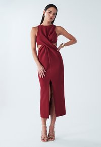 Czerwona sukienka bez rękawów z ozdobnym skręceniem na przodzie i wycięciami po bokach. Posiada rozcięcie sięgające uda oraz dopasowany krój.