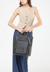 Donna con un gilet senza maniche beige e pantaloni blu, che tiene una borsa a tracolla in pelle nera con una tasca frontale con zip e un'etichetta che legge "IO SONO UNA BORSA RESPONSABILE."