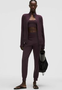 Bordo sporta komplekts, kas sastāv no ar rāvējslēdzēju aizveramas jakas, sporta krūštura un šaurām biksēm. Gluda auduma ar pieguļošu krāsu. Melns jogas paklājiņš rokā.