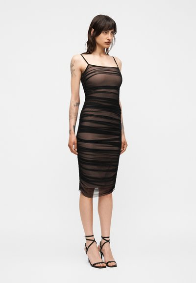 Γυναίκα που φοράει μαύρο διάφανο μίντι φόρεμα με οριζόντιες ραβδώσεις και λεπτές τιράντες, συνδυασμένο με μαύρα σανδάλια με λεπτά λουράκια και ψηλό τακούνι.