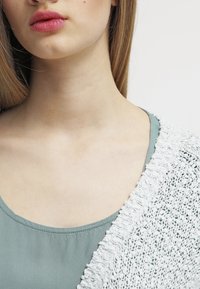 Lichtgroene top met een ronde hals, gelaagd onder een witte, ingewikkeld gehaakte cardigan die een textuurontwerp laat zien.